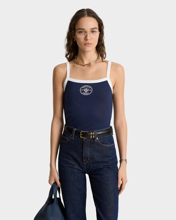 Woman wearing glasses, dark jeans, holding a blue bag, and dressed in the Eden Crest Embroidered Pique Tank in navy with white trim.
