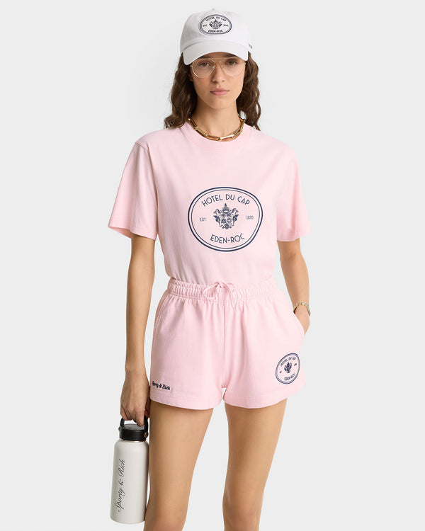 Woman in the Eden Crest T-Shirt - Pink Dolphin/Navy and shorts, white cap, holding a water bottle against a plain background. Made from 100% cotton in Portugal for comfort and style.
