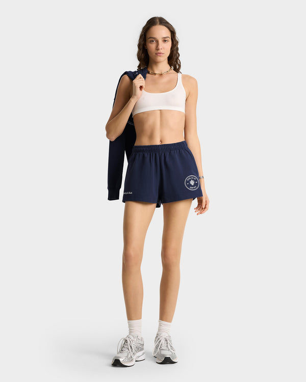 Woman in white sports bra, Eden Crest Disco Short - Navy/White, and sneakers stands holding a navy jacket over her shoulder.