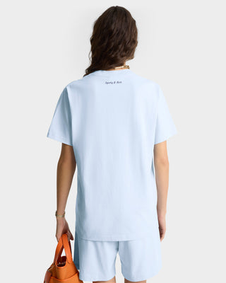 Person shown from the back against a plain background, wearing the Eden Crest T-Shirt - Ciel/Navy (100% cotton, made in Portugal), styled oversized with shorts and holding an orange bag.