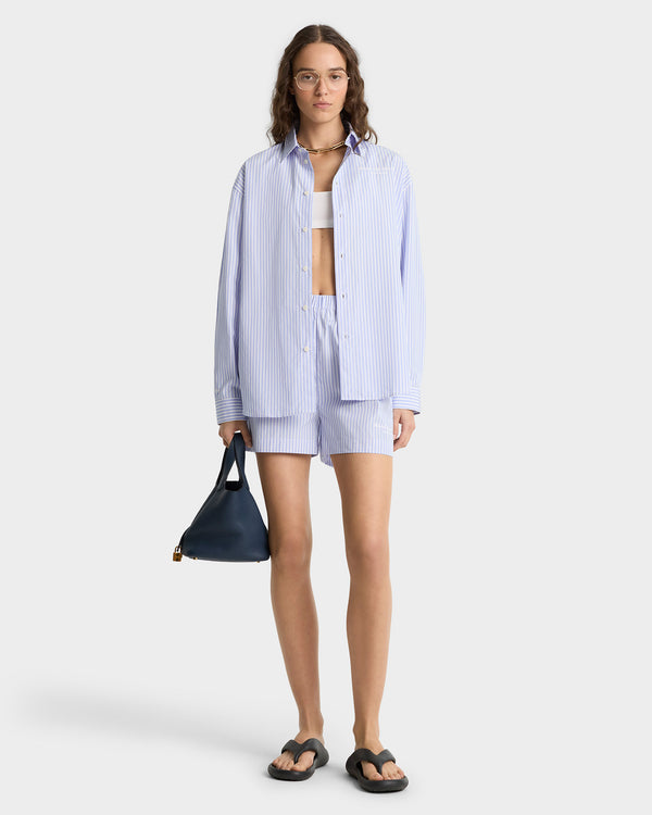 Woman wearing the Eden Script Oversized Shirt in blue/white stripe, styled with matching shorts, a white crop top, sandals, and a black handbag.