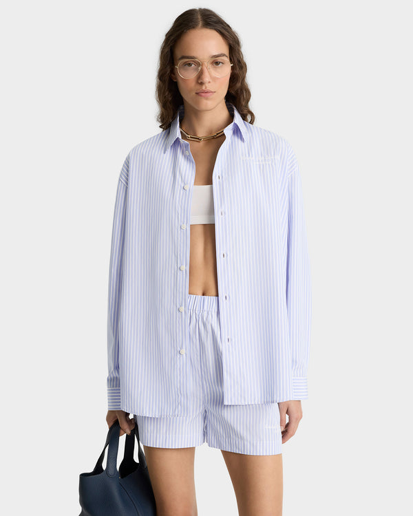 Woman wearing the Eden Script Oversized Shirt in blue and white stripes with matching shorts, glasses, and a navy bag, standing against a plain background.
