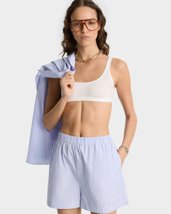 A woman in glasses wears a white crop top with Eden Script Poplin Shorts in blue/white stripe, made in Portugal, holding the matching shirt over her shoulder.