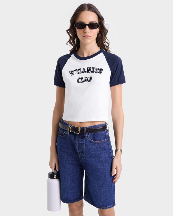 A woman in sunglasses and denim shorts wears the Wellness Club Flocked Baby Tee in White/Dark Navy, crafted from cotton rib fabric, and holds a white water bottle.