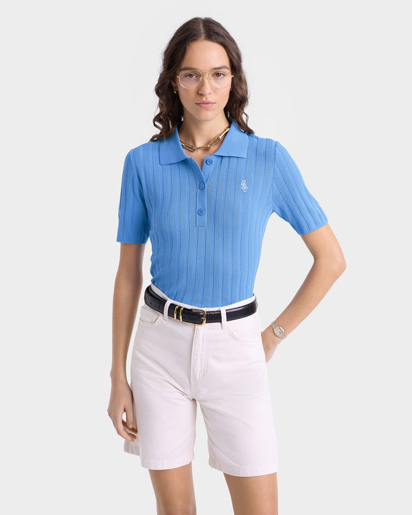 Woman wearing the SRC Ribbed Polo in Juniper/White with a buttoned collar and embroidered logo, paired with white shorts, glasses, and jewelry, standing against a plain background.