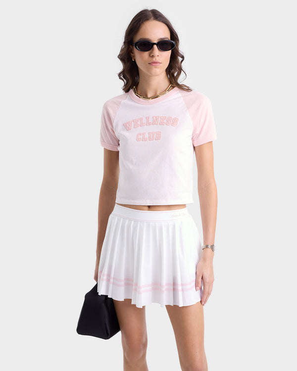 Woman in sunglasses wears the Wellness Club Flocked Baby Tee in White/Pink Dolphin, styled cropped with a white pleated skirt and black bag.