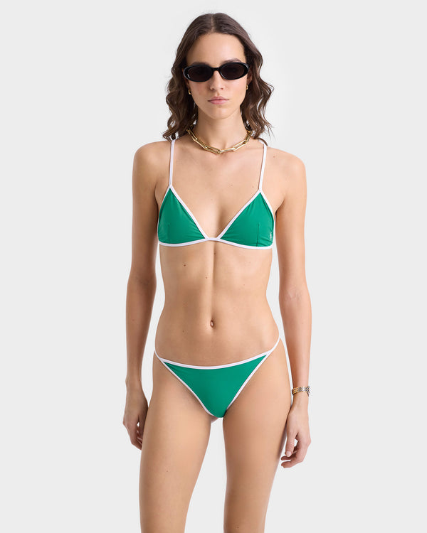A woman in the Kate Bikini Top - Spring Green/White, sunglasses, and a gold necklace—crafted from recycled ECONYL® Nylon—stands against a plain background.