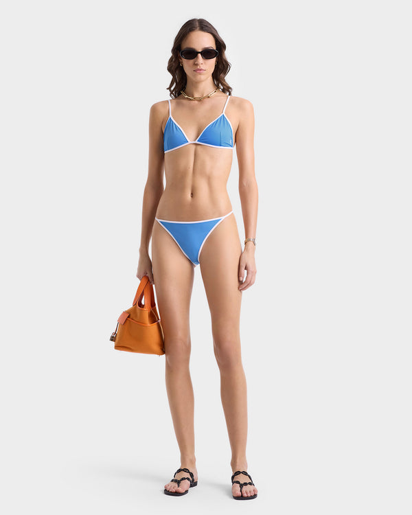 Woman wearing the Kate Bikini Bottom in Juniper/White with white trim, sunglasses, black sandals, and an orange handbag, standing against a white background.