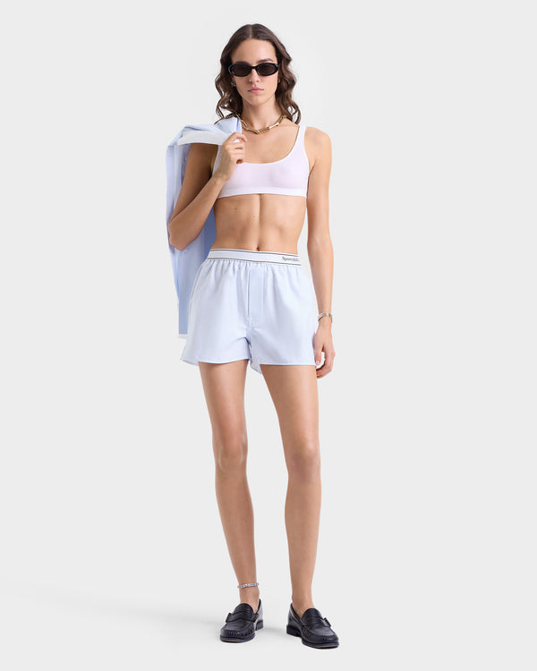 A woman in sunglasses wears a light bralette, loafers, and the Serif Logo Oxford Boxer in blue/white thin stripes with an elastic waistband, holding a jacket over her shoulder.
