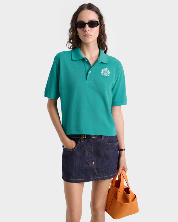 Woman wears the H&W Crest Pique Polo in Spring Green/White with an embroidered logo, paired with a denim skirt, sunglasses, and an orange handbag against a white background.