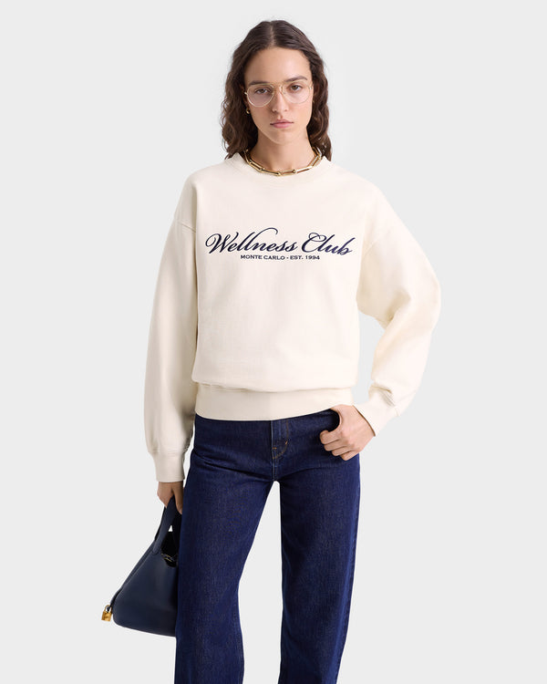 Woman in glasses wearing the Wellness Club Embroidered Crewneck in Cream/Navy with jeans, holding a black handbag.