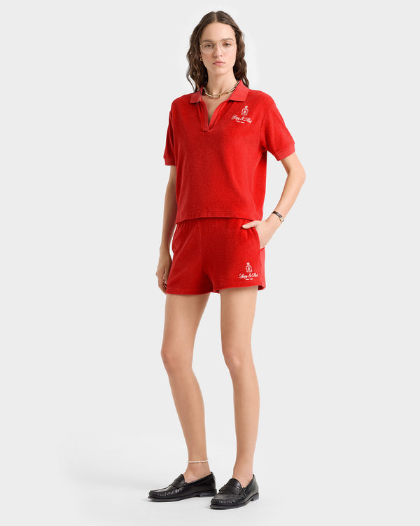 A woman stands on a white background wearing the Vendome Terry Short in fire red/white, paired with a matching short-sleeve polo and black loafers.