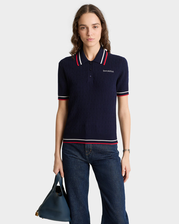 A woman in a Serif Logo Mini Cable Polo in navy, white, and ruby with an embroidered logo holds a blue handbag and wears blue jeans.