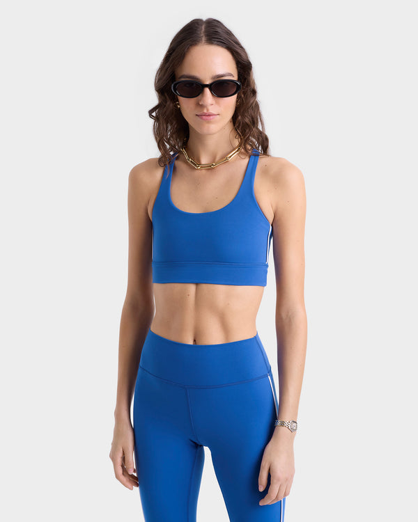 Close up of model wearing bue sports bra with thin racerback straps and subtle white side stripes, standing up in a light gray background.