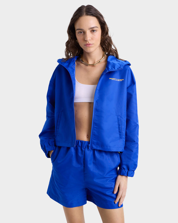 Close up view of model wearing blue nylon full-zip jacket with a high collar, featuring gold 'Sporty & Rich' text on the chest, side pockets, and elasticated cuffs, paired with matching shorts.