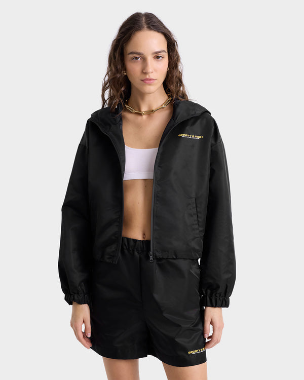 Model wearing black nylon full-zip jacket with a high collar, featuring gold 'Sporty & Rich' text on the chest, side pockets, and elasticated cuffs, paired with matching shorts.