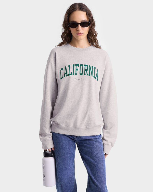 Model wearing heather grey crewneck sweatshirt with green 'CALIFORNIA' text across the chest and smaller 'Sporty & Rich' text below, featuring ribbed hem and cuffs.