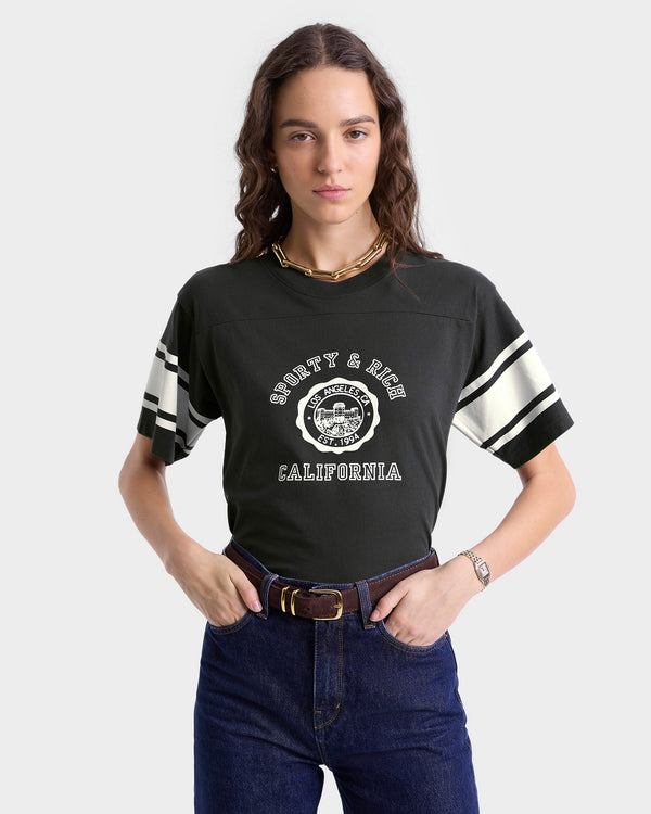 Close up of model wearing faded black T-shirt with double white stripes on each sleeve and a white circular 'Sporty & Rich Los Angeles Est. 1994' graphic on the chest, featuring a crew neckline and short sleeves.