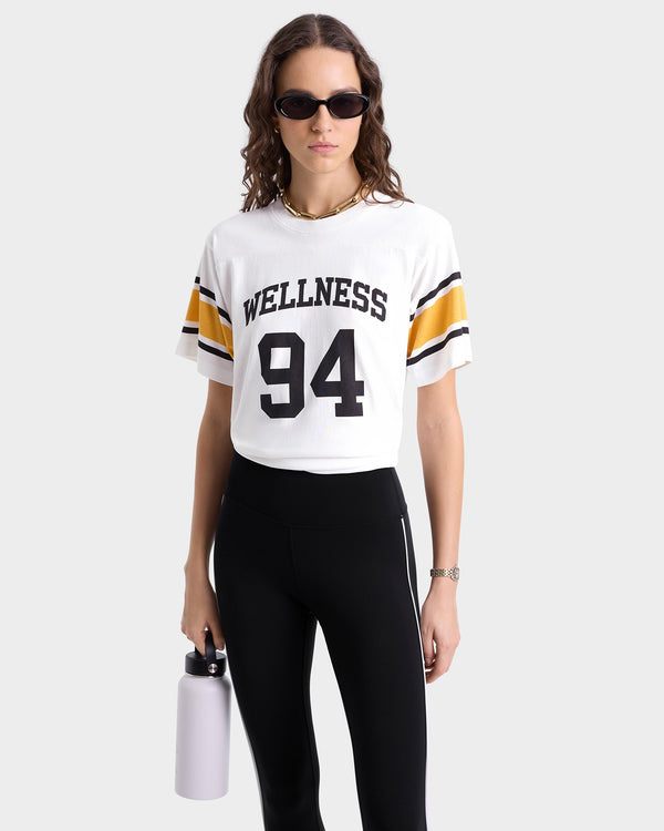 Close up of model wearing white jersey-style T-shirt with 'WELLNESS 94' in bold black lettering on the front, featuring gold and black stripes on each sleeve and a crew neckline.