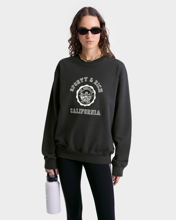 Close up of model wearing faded black crewneck sweatshirt with a white circular 'Sporty & Rich Los Angeles CA Est. 1994' graphic on the chest, featuring ribbed hem and cuffs.