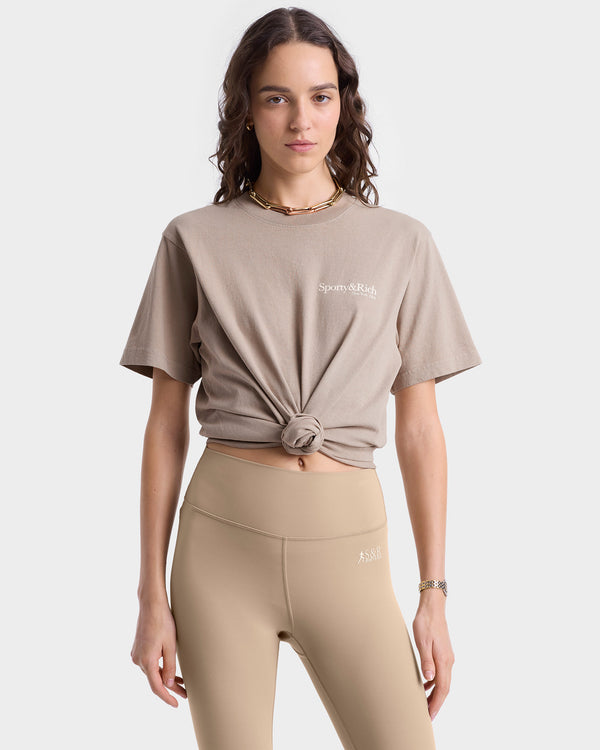 Close up of model wearing tan T-shirt with white 'Sporty & Rich' text on the left chest, featuring a crew neckline and short sleeves.