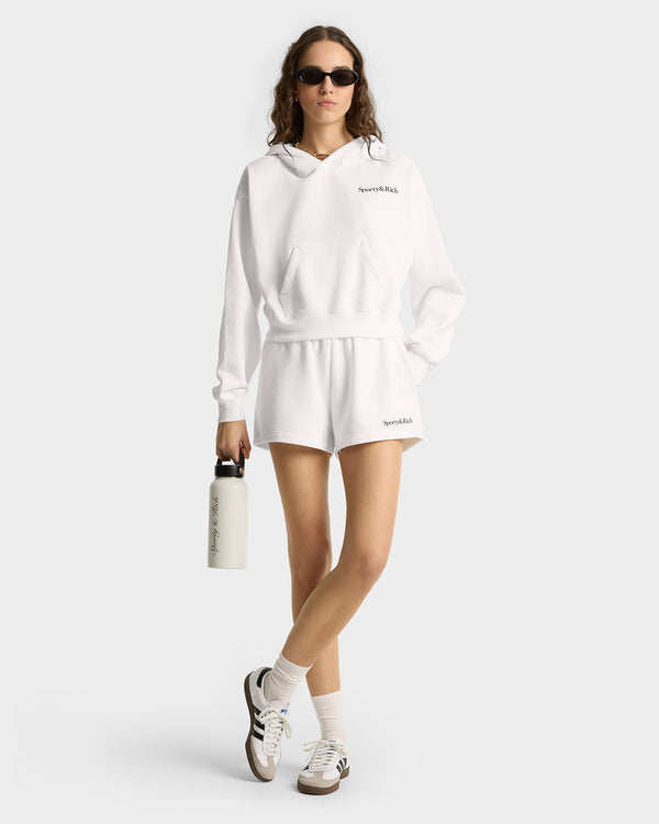 Woman in white Serif Logo Soft Disco Short - White/Navy and hoodie, wearing sunglasses and holding a water bottle, stands against a light background.