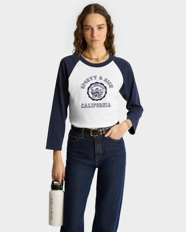 Woman in the California Emblem Soft Baseball Tee in white/navy, made from 100% cotton in Portugal, paired with jeans and holding a white water bottle.