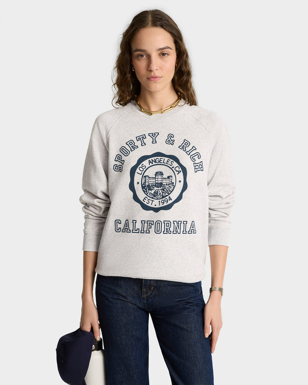 Young woman in a heather gray California Emblem Soft Crewneck and jeans, holding a navy cap, stands facing forward.