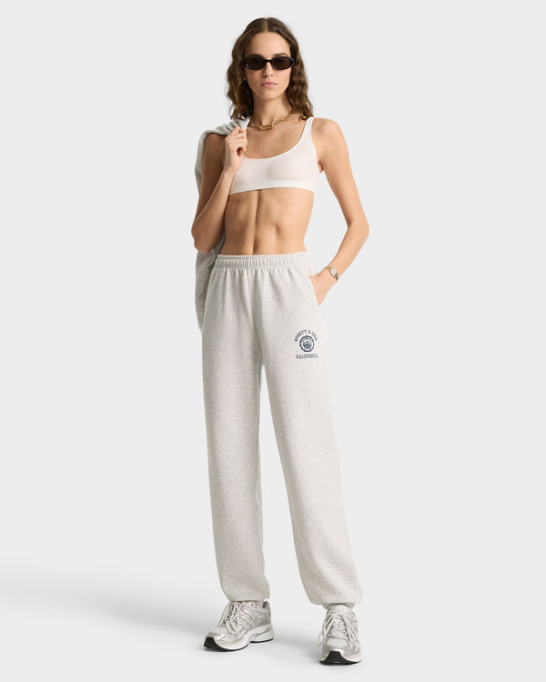 Woman in sunglasses wears a white sports bra, sneakers, carries a bag over her shoulder, and sports the California Emblem Soft Sweatpant in Heather Gray/Navy.