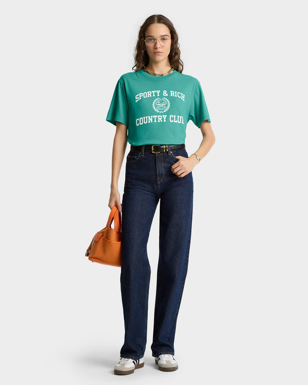 Woman wearing a green Varsity Crest Soft T-Shirt - Spring Green/White, jeans, glasses, and holding an orange handbag.