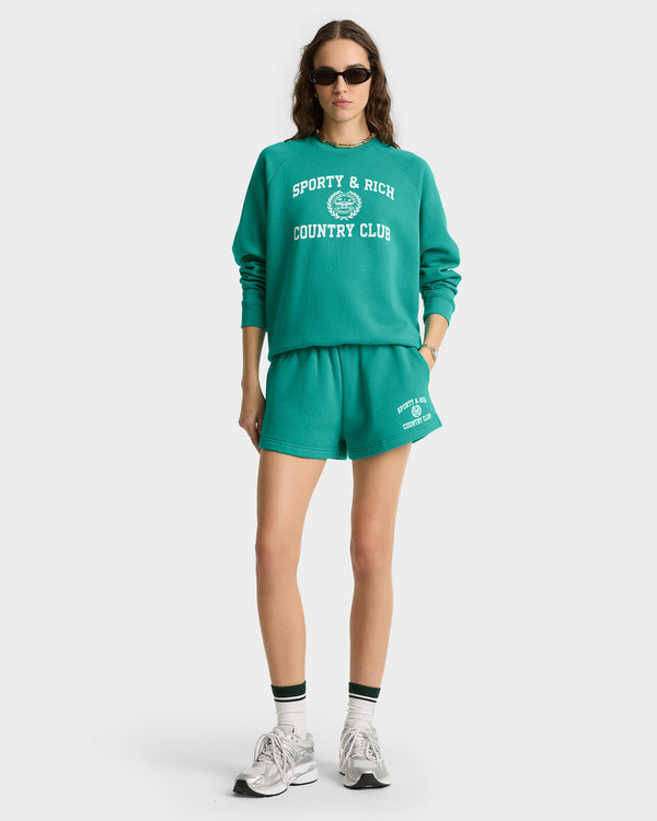 Woman in a pre-shrunk Varsity Crest Soft Crewneck in spring green/white, paired with shorts, sunglasses, and sneakers.