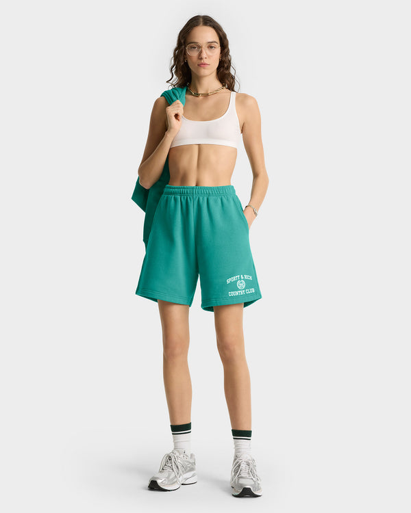 Woman in Varsity Crest Soft Gym Short – Spring Green/White with elastic waistband, white sports bra, and sneakers stands holding a matching jacket over her shoulder.