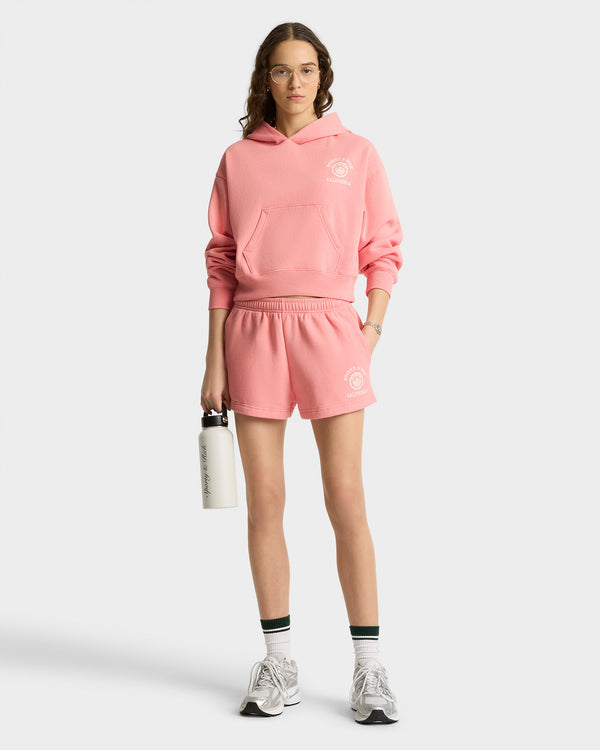 Woman in a pink hoodie and California Emblem Soft Disco Short - Carnation/White, holding a water bottle, wearing sneakers, glasses, and striped socks.