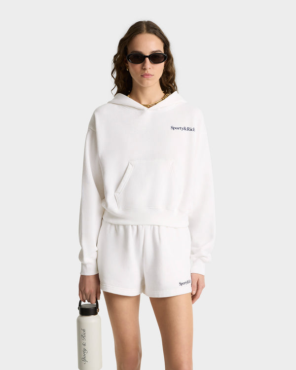 Woman wearing the Health Is Wealth Soft Cropped Hoodie in White/Navy, paired with shorts and sunglasses, holding a water bottle against a plain background.