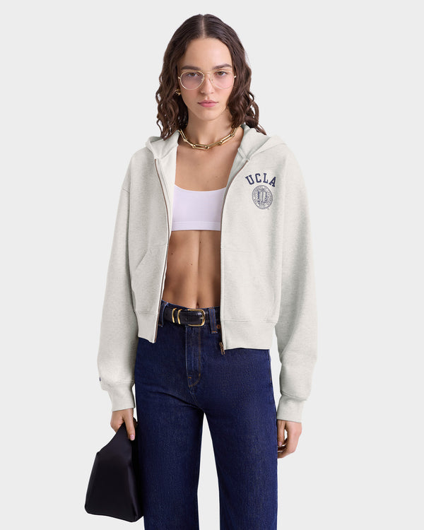 Woman in glasses wears a UCLA Crest Cropped Zip Hoodie - Heather Gray/Navy, a white crop top, and blue jeans, holding a black bag against a plain background.