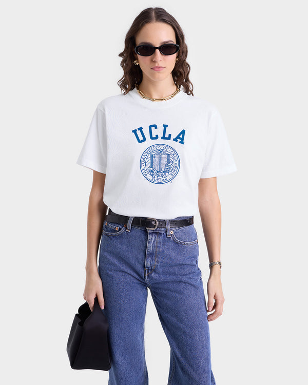 A woman wearing sunglasses, blue jeans, and holding a black handbag poses against a plain background in the UCLA Crest T-Shirt - White/Blue, made from USA grown cotton.