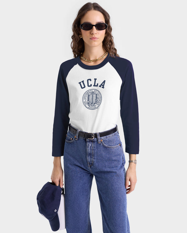 Woman wearing sunglasses, jeans, and the UCLA Crest Baseball Tee - White/Navy, holding a navy cap against a plain light background. Made in the USA.