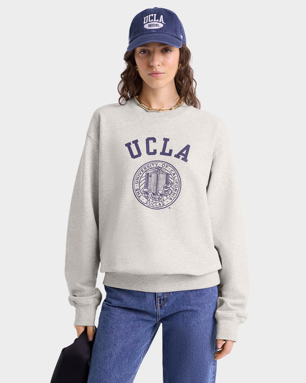 Woman wearing the UCLA Crest Crewneck in Heather Gray/Navy, blue jeans, and a UCLA cap, holding a dark item by her side.