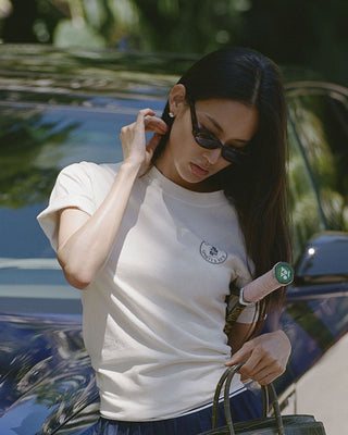 A woman wearing a white short-sleeve crewneck t-shirt with a small circular logo on the left chest, styled with dark sunglasses, holding a green handbag, and standing next to a blue car outdoors.