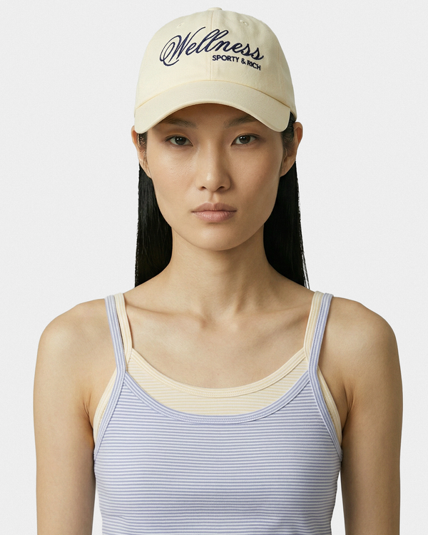Woman wearing a beige 'Wellness' cap and striped tank top on a white background