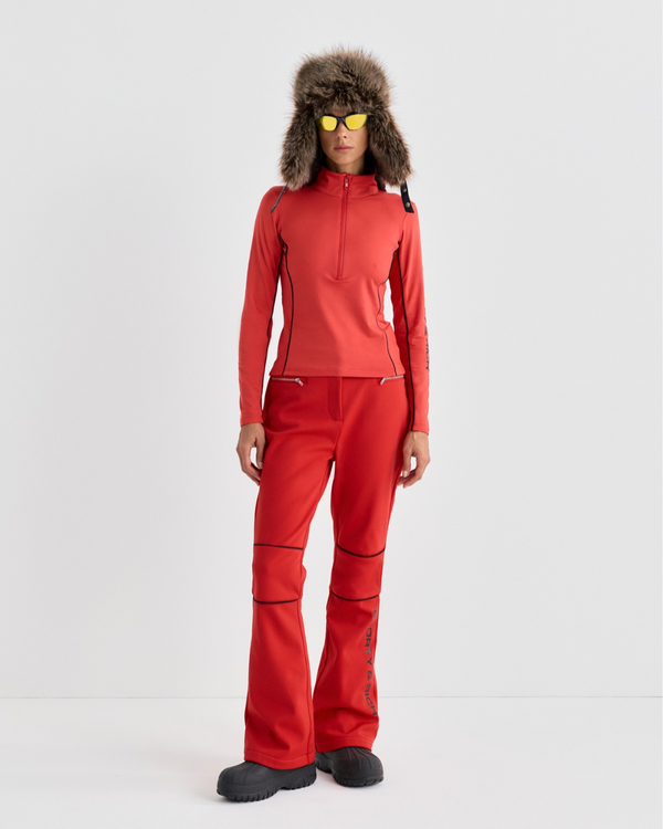 A person in a red zip-up top, Boso Bold Logo Ski Pants - Rosso, a fur-lined trapper hat, and yellow-tinted sunglasses.