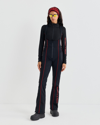 A person wears a black Divy Bold Logo Ski Jumpsuit with red piping, a red headband, and yellow sunglasses.