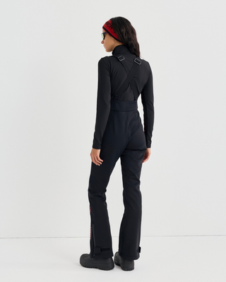 A person wears a black Divy Bold Logo Ski Jumpsuit with red piping, a red headband, and yellow sunglasses.