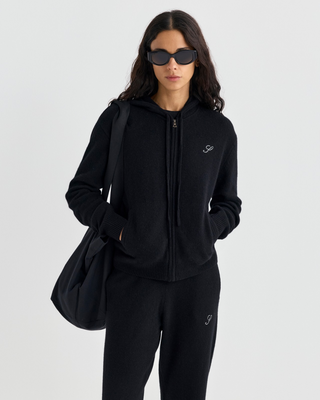 A person in a black zip-up hoodie and Ivana S Logo Cashmere Trousers, carrying a large black tote bag.