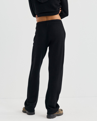 A person in a black zip-up hoodie and Ivana S Logo Cashmere Trousers, carrying a large black tote bag.