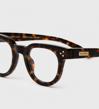 Close-up of a pair of round tortoiseshell eyeglass frames on a clean white background, showing the thick acetate pattern and a small gold-tone brand plaque on the temple.