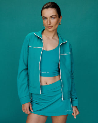 Woman wearing the Court Jacket - Teal, styled with a crop top and skirt, standing against a matching teal background.