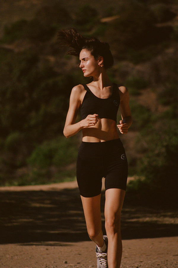A woman jogs outdoors on a sunny, tree-lined dirt path wearing the Biker Short N.02 - Black from Sporty & Rich.