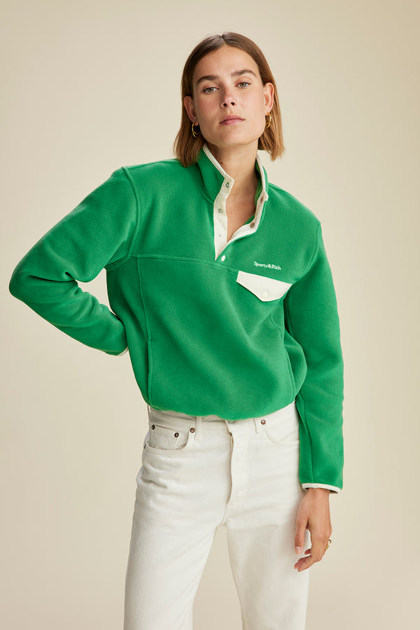 A woman models the Buttoned Polar Sweatshirt in Kelly/Cream with snap button closure, paired with white jeans and posed against a beige background—an ideal choice for stylish winter wear.