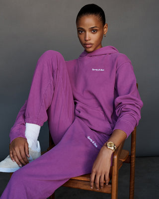 A woman wearing the Classic Logo Hoodie - Magenta, made from 100% cotton, sits on a chair against a gray background. She pairs it with matching tracksuit bottoms, white sneakers, and a watch.